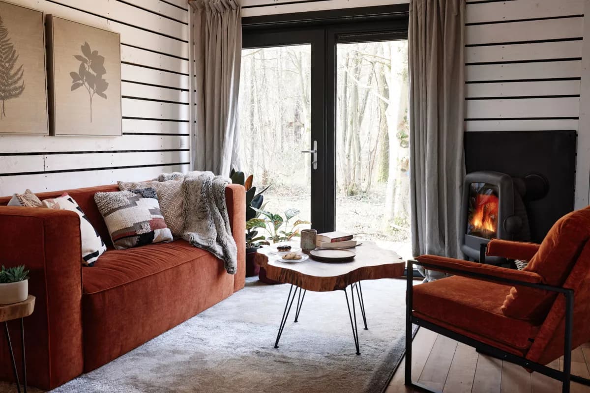 Le salon : canapé et fauteuil rouille, table basse en bois brut et cheminée à bois allumée, porte-fenêtre ouverte sur la forêt